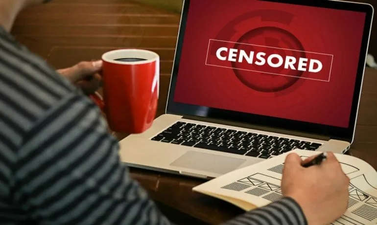 Мужчина с кружкой кофе в руке смотрит на экран ноутбука с уведомлением о цензуре контента